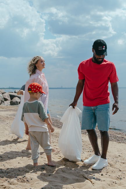 A group of three people, including a man and two children, are participating in a beach clean-up activity. The man, dressed in a red T-shirt, light blue denim shorts with some frayed edges, and a blac
