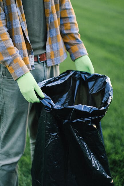 A group of three people, including a man and two children, are participating in a beach clean-up activity. The man, dressed in a red T-shirt, light blue denim shorts with some frayed edges, and a blac